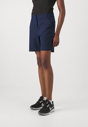 Person iført marineblå shorts, sort ærmeløs top, hvide sokker og sorte Nike-sneakers, der står på en ensfarvet hvid baggrund.