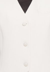 Blusa blanca de botones con diseño de escote en V, que cuenta con cuatro botones circulares y un pequeño bolsillo en el pecho. Textura de tela suave.