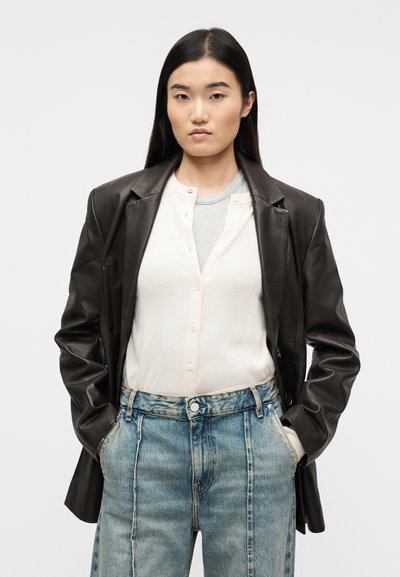 Νεαρή γυναίκα με μακριά σκούρα μαλλιά που φοράει μαύρο δερμάτινο σακάκι, ανοιχτό αμάνικο με κουμπιά πάνω από γκρι μπλούζα και μπλε τζιν με ψηλή μέση.