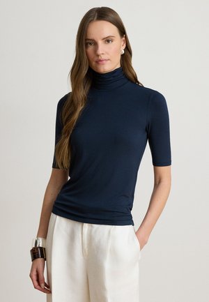 Femme aux longs cheveux bruns portant un haut à col roulé bleu marine à manches courtes et un pantalon blanc, debout avec une main dans la poche.