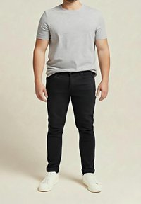 Homme portant un t-shirt à manches courtes gris clair, un pantalon noir ajusté et des baskets blanches, debout devant un fond beige uni.