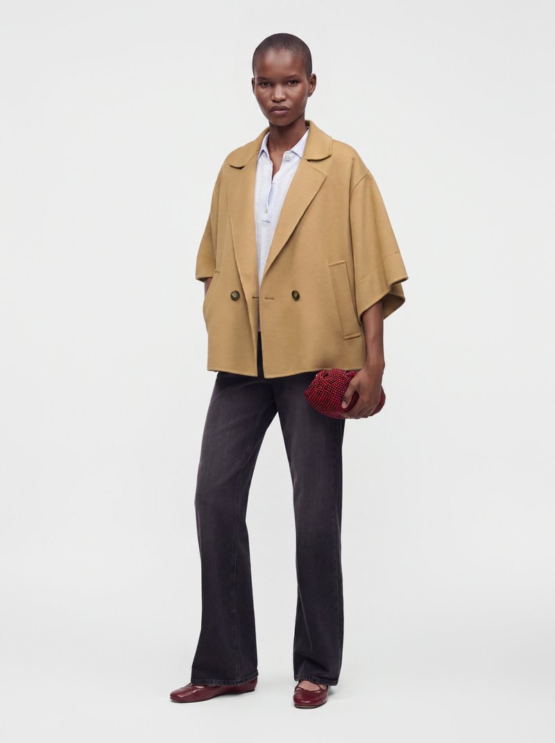 Femme portant une veste oversize camel, un chemisier blanc, un jean large foncé, des chaussures rouges et tenant une petite pochette rouge perlée.