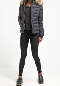 Černá péřová bunda s umělou kožešinou na kapuci, se zipem a designem ve tvaru diamantů. Nositelná s černými legínami a lesklými černými botami.