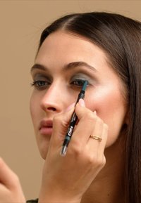 Svart eyeliner blyant i hånden, påfører en mørk teal nyanse på øyelokket. Modellen har glatt hud, rett brunt hår, og nøytral sminke.