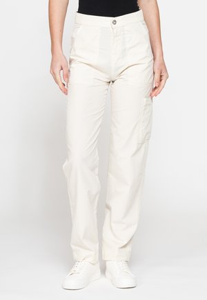 Persona che indossa pantaloni cargo bianchi, top nero e sneakers bianche, in piedi davanti a uno sfondo bianco semplice.