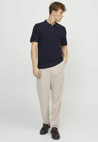 Polo blu navy a maniche corte con finitura testurizzata, abbinato a pantaloni beige plissettati e scarpe in pelle marroni con dettagli decorativi.