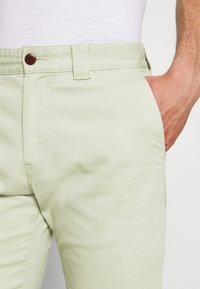 Pantaloni di cotone verde chiaro con una texture morbida, dotati di chiusura con bottone marrone e tasche laterali. Vestibilità su misura con dettagli di cucitura sottili.
