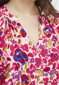 Blus med blommönster som har en vit bakgrund med livfulla rosa, röda, blå och gröna blommor, samt en knappad front med en plisserad halsringning.