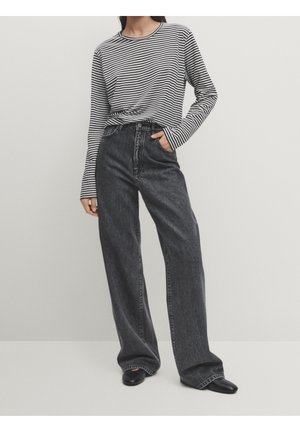 Personne portant une chemise à manches longues rayée noir et blanc, rentrée dans un jean noir délavé taille haute à jambes larges, avec des chaussures noires.