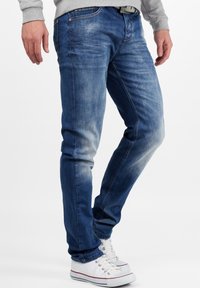 Jean en denim bleu au design ajusté, avec des décolorations légères, un style classique à cinq poches et des coutures visibles. Porté avec des baskets blanches.