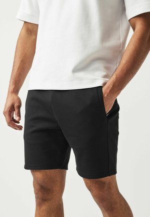 Homme portant un short noir au niveau des genoux et un t-shirt blanc avec une main dans la poche, sur un fond uni.