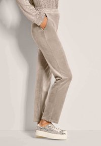 Persoon in beige velours broek met zijzakken en sneakers met slangenprint op het platform, staand tegen een lichte muur.