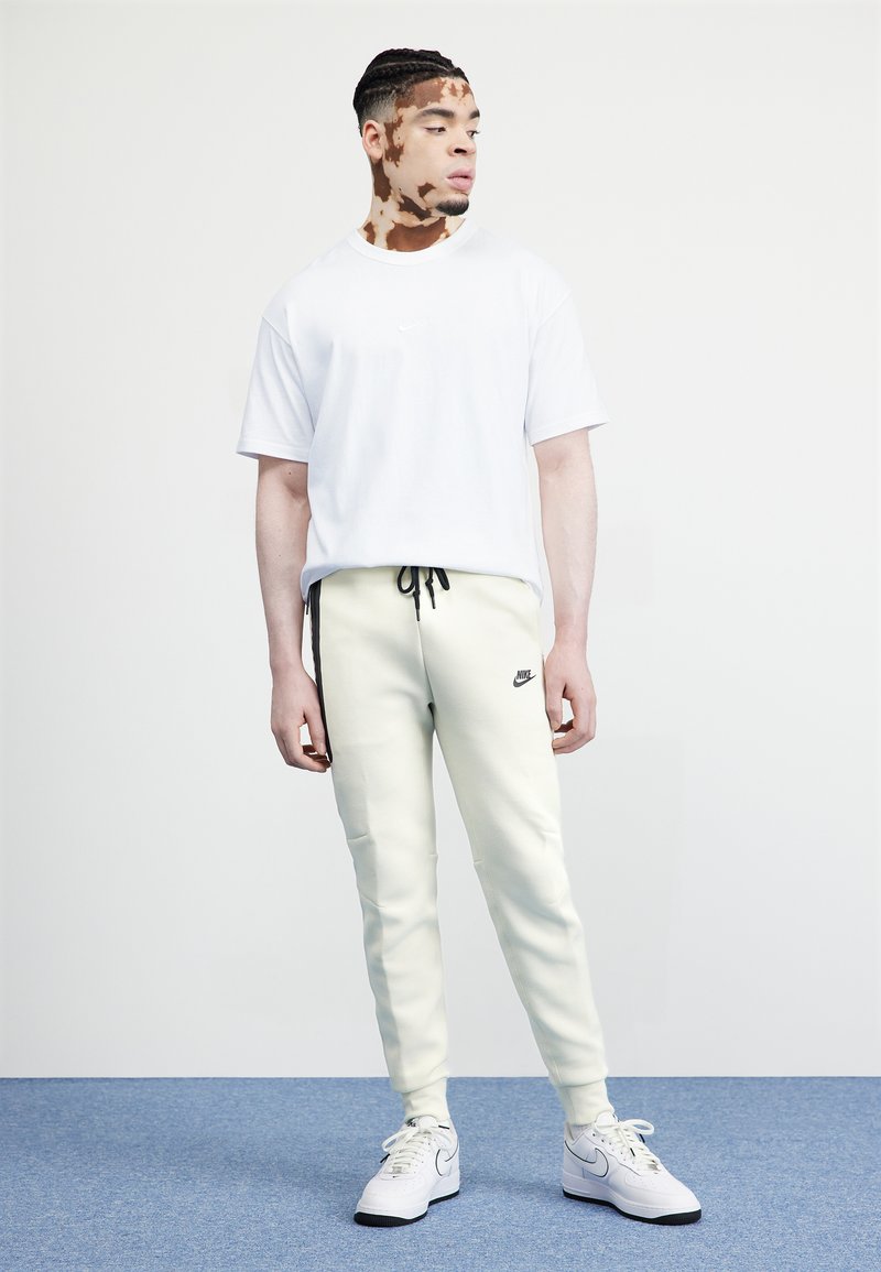 Weißes Baumwoll-T-Shirt mit kurzen Ärmeln, kombiniert mit hellgelben Sweatpants, die ein schwarzes Nike-Logo zeigen, abgerundet mit weißen Turnschuhen.