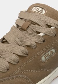 Brown suede sneaker with flat beige laces, Fila logo tag, and textured side panel. Features rounded toe and rubber outsole.
