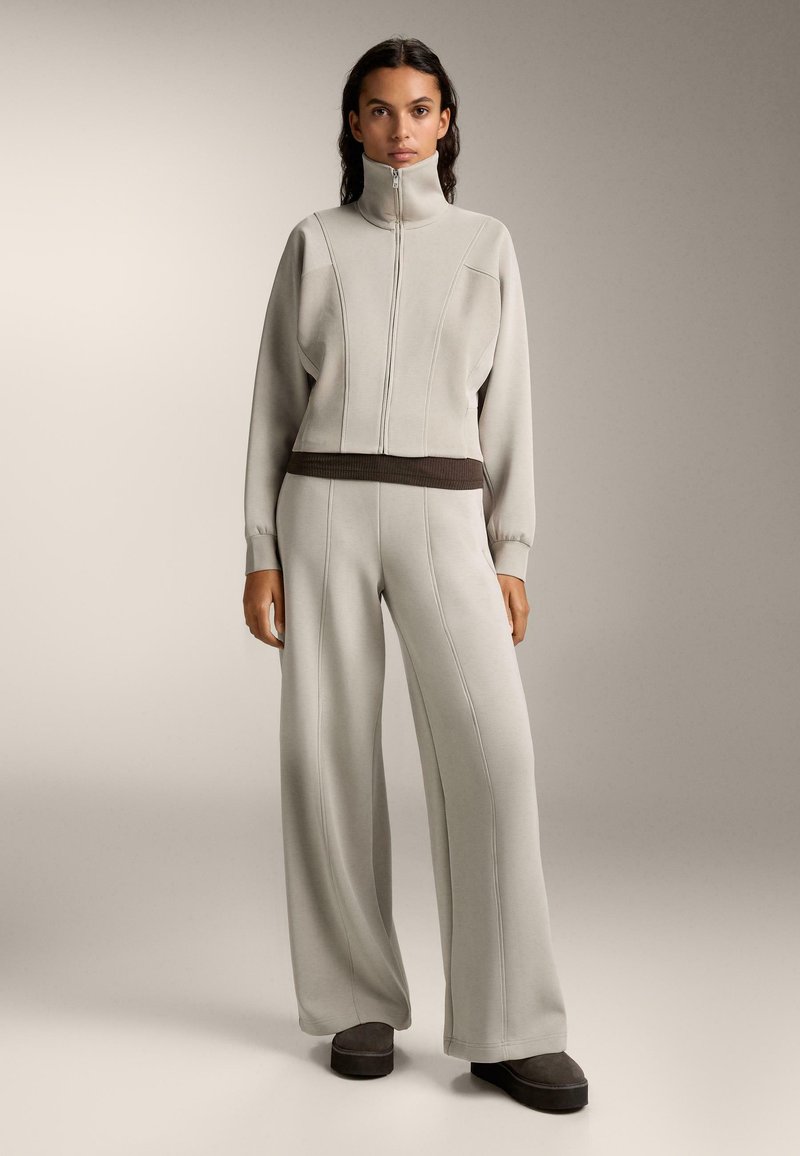 Svetlo šedý teplákový komplet s vysokým golierom a zipovým predným zapínaním. Obsahuje rebrované akcenty a široké nohavice. Čierne akcenty v pase a na obuvi.