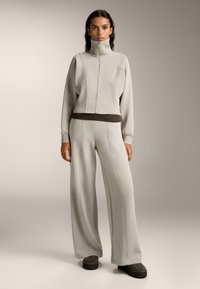 Svetle šedý teplákový komplet s vysokým límcem a zipem vpředu. Obsahuje žebrované detaily a kalhoty širokého střihu. Černé akcenty na pase a obuvi.