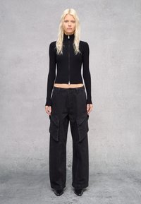 Camiseta negra de manga larga con cremallera y canalé, combinada con pantalones cargo negros de pierna ancha que presentan bolsillos laterales y una textura suave. Zapatos de tacón negros.