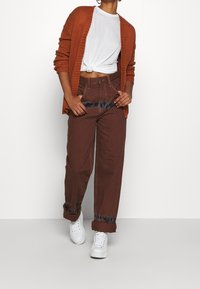 Pantalon marron taille haute avec des accents tie-dye bleus aux poignets, associé à un t-shirt blanc noué et un cardigan orange. Baskets blanches.