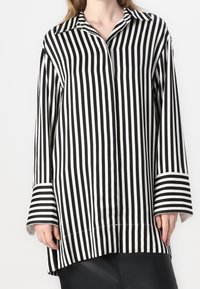 Camiseta de manga larga a rayas en blanco y negro con cuello y mangas anchas. Textura suave; las rayas varían en ancho. El dobladillo inferior es ligeramente más largo.