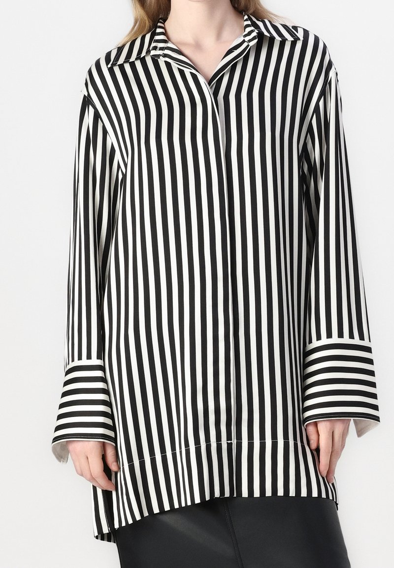 Camicia a maniche lunghe a righe bianche e nere con colletto e maniche larghe. Tessuto liscio; le righe variano in larghezza. L'orlo inferiore è leggermente più lungo.