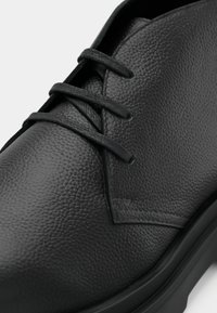 Μαύρο δερμάτινο παπούτσι με υφή, κλειστό σύστημα κορδονιών και στρογγυλή μύτη. Η σόλα είναι επίπεδη και λίγο ανυψωμένη.