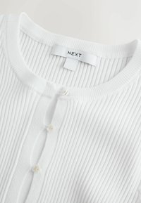 Fehér bordázott kötött kardigán kerek nyakkivágással és apró, virág alakú gombokkal, a nyakrésznél „NEXT” címkével ellátva.