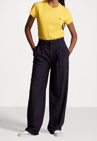 Osoba v žlutém tričku s krátkým rukávem, černých širokých kalhotách s rukama v kapsách a bílých teniskách na jednobarevném pozadí.