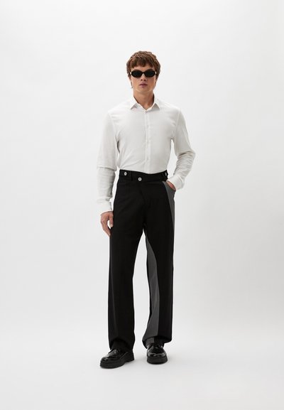 Weißes Langarmhemd, kombiniert mit weiten, schwarz-grauen Hosen mit Knopfakzenten und glatter Textur. Das Model trägt Sonnenbrillen und Schuhe.