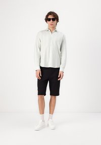 Chemise à manches longues rayée vert clair, shorts noirs, baskets blanches et lunettes de soleil noires. Tissu lisse avec une coupe décontractée et des shorts jusqu'aux genoux.
