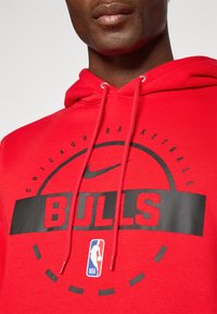 Nærbilde av en person som har på seg en rød Chicago Bulls basketball-hettegenser med svart tekst og NBA-logo på brystet.