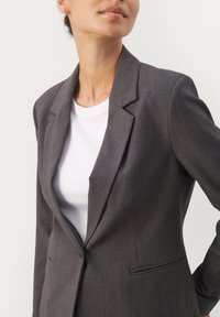 Šedý přizpůsobený blazer s jedním knoflíkem, s klopami a dvěma bočními kapsami, vrstvený přes bílý žebrovaný top.