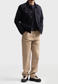 Chaqueta de denim negra con desgaste, camiseta oscura, pantalones cargo beige y zapatillas negras y crema. Presenta bolsillos frontales y un cinturón.