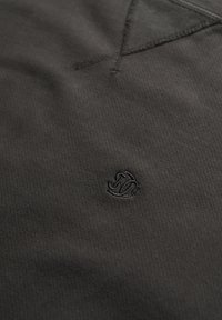Tela negra texturizada con un logo sutil bordado en hilo oscuro. Presenta un diseño de cuello en V y costuras bien elaboradas. Apariencia simple y moderna.