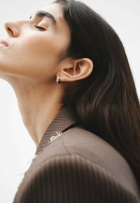 Pendiente de aro plateado en una modelo, con cabello largo y oscuro, que lleva una chaqueta marrón a rayas. Enfoque en el detalle del pendiente y la textura suave de la piel.