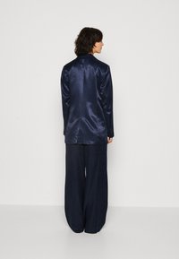 Costum din satin de culoare albastru marin, compus dintr-un sacou lejer cu ventilație la spate și pantaloni evazați, având o textură netedă și lucioasă.