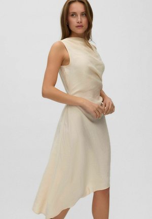 Jeune femme portant une robe crème sans manches, longueur genou, avec un ourlet asymétrique, debout devant un fond clair uni.