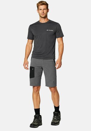 Mann trägt dunkelgraues Columbia T-Shirt, graue knielange Shorts mit schwarzer Tasche, schwarze Socken und Wanderschuhe, steht vor weißem Hintergrund.