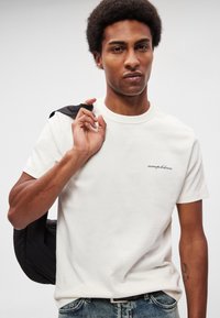 Biele bavlnené tričko s krátkym rukávom a okrúhlym výstrihom. Na hrudi má malú čiernu vyšívanú značku. Nosené s džínsami.