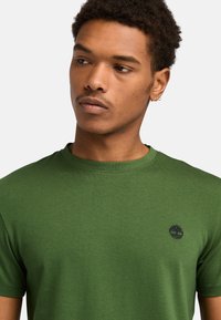 Grøn bomulds T-shirt med rund halsudskæring. Har et lille sort logo på brystet. Glat tekstur, korte ærmer og en tætsiddende pasform.