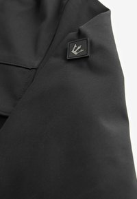 Material negru cu o textură netedă și finisaj mat; include o etichetă pătrată cu un emblema argintie în colț.