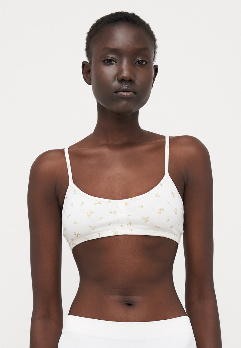 White bralette with yellow floral print, thin straps, and button detail at the front. Soft, stretchy fabric.
