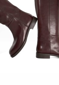 Burgundy leather knee-high boots with a smooth finish, rounded toe, and low, solid black heel, featuring minimalistic design details.