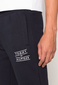 Marineblauwe sweatpants met een zachte textuur, voorzien van een wit "TOMMY HILFIGER" logo en een klein rood en blauw embleem. Inclusief zijzakken.