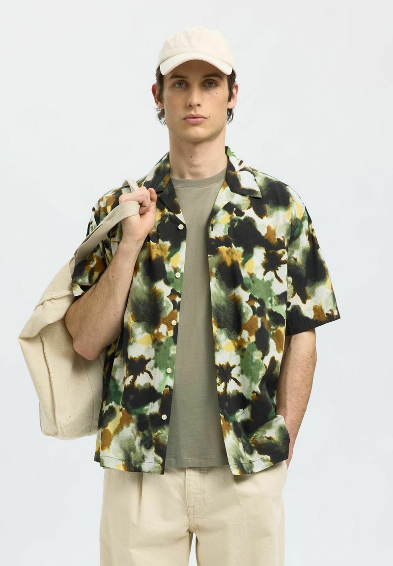Kurzarmhemd in einem mehrfarbigen Camouflage-Muster mit Tönen von Grün, Schwarz und Braun, kombiniert mit einem hellen T-Shirt und cremefarbenen Hosen.