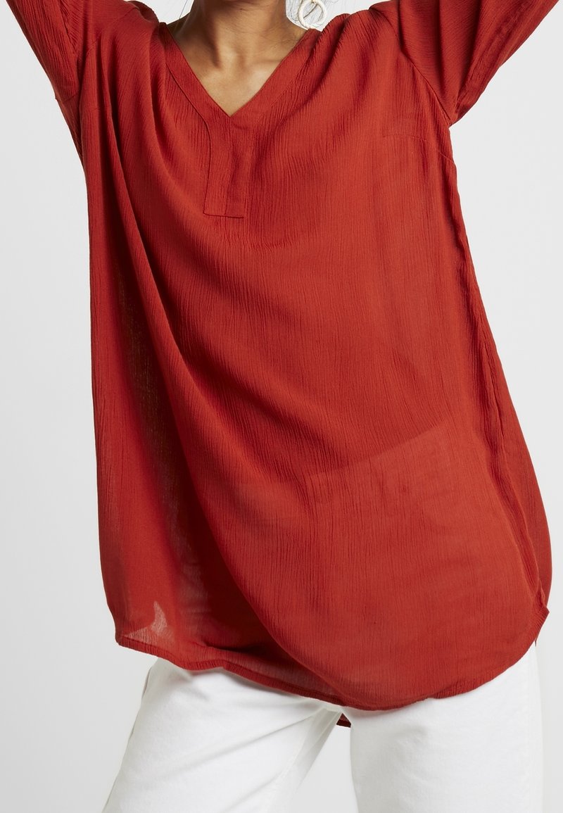 Blouse rouge texturée avec un col en V et des manches longues. Coupe ample et aspect légèrement transparent. Portée sur un pantalon blanc.