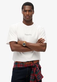 Camiseta blanca de algodón con el logo de "Superdry". El modelo lleva un reloj de cuero marrón y tiene una camisa de cuadros roja y verde anudada a la cintura. Jeans oscuros.