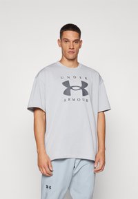 Lys grå oversized T-skjorte med rund hals. Har svart "UNDER ARMOUR"-tekst og logo på forsiden. Laget av en bomullsblanding.