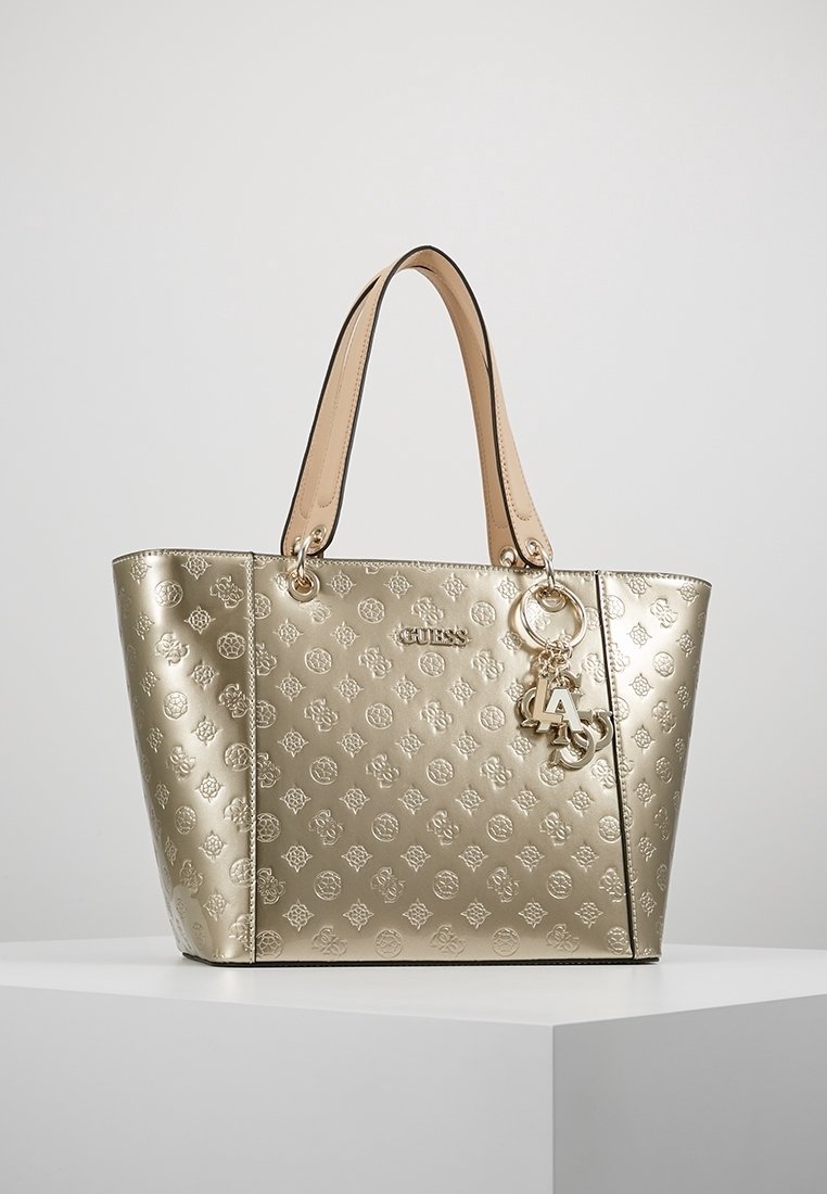 Metalická zlatá taška přes rameno s embosovaným květinovým vzorem, hnědými popruhy a stříbrným přívěskem. Prostorná s pevným tvarem a otevřeným vrškem.