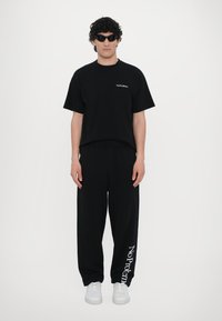 Svart bomullst-skjorte med teksten "No Problemo", kombinert med svarte joggebukser som har hvit tekst på siden, fullført med hvite sneakers.