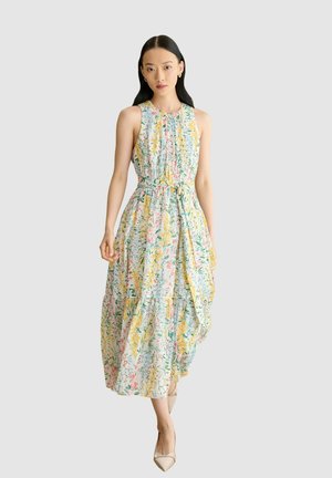 Femme portant une robe midi sans manches à fleurs avec une taille nouée et un corsage plissé, associée à des chaussures pointues beiges sur un fond uni.